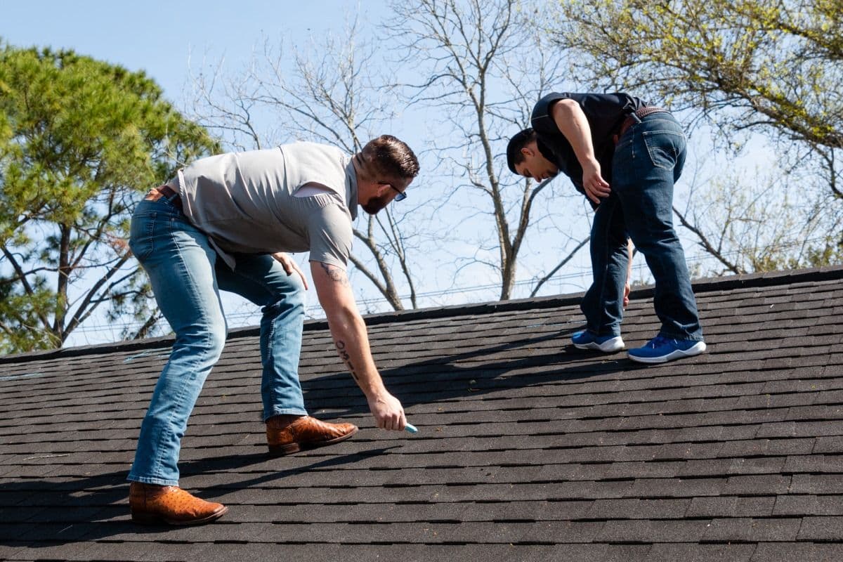 Two men inspecting a roof, one marking it while the other observes, on a sunny day.