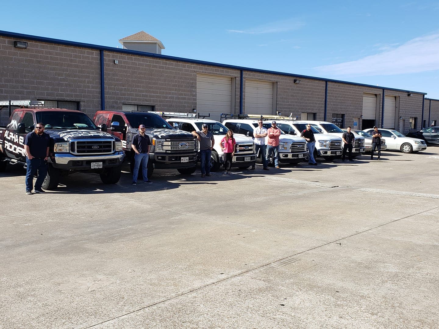 Group of professionals posing with company trucks outside a warehouse on a sunny day.